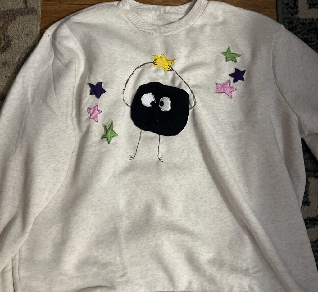 Grey swetshirt with appliqué "soot sprite" holding up a yellow star, surrounded by 6 other little multi-colored stars (inspired by Spirited Away).